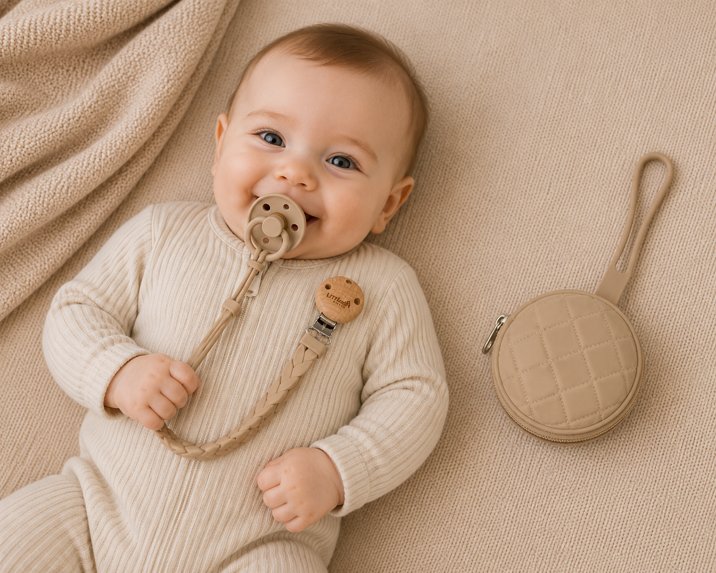 Baby lying on a beige blanket with a pacifier and teething ring, next to a matching dummy case.