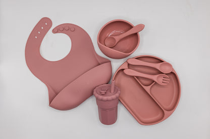 Powder rose silicone feeding set flat lay, including bib, divided tray, cup, and utensils