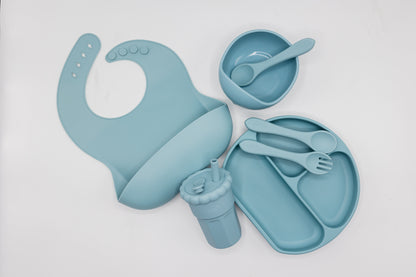 Dusty blue silicone feeding set flat lay, with bib, divided plate, cup, and utensils