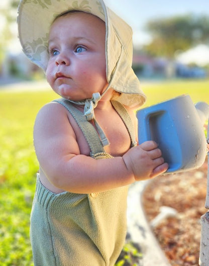 Kids watering can toy with light blue cup; child in green overalls and sun hat
