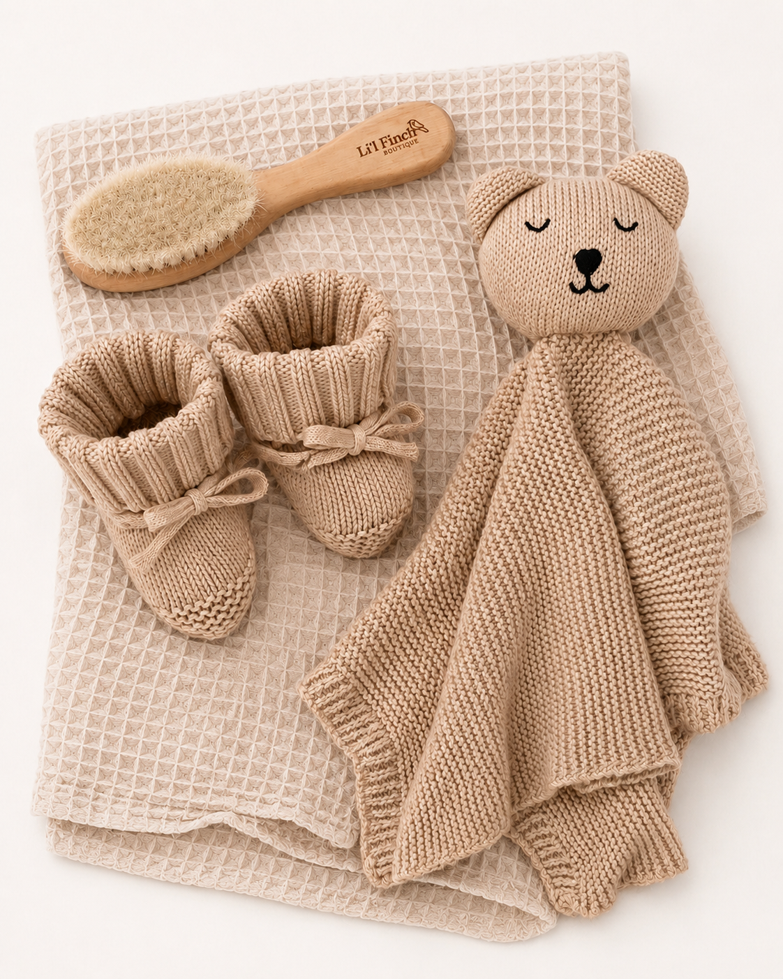 Baby items including a knitted bear comfort toy, booties, and a brush on a textured fabric background.